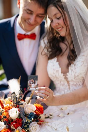 Newlyweds share intimate moment with autumn-themed wedding bouquet featuring red and white flowers and blue accents.