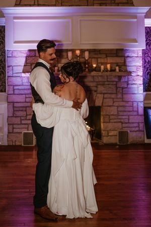 A couple shares an intimate first dance together in a ballroom with purple uplighting and a stone fireplace.