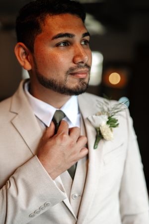 Close-up view of groom in white suit and boutonniere adjusts his tie while looking off camera.