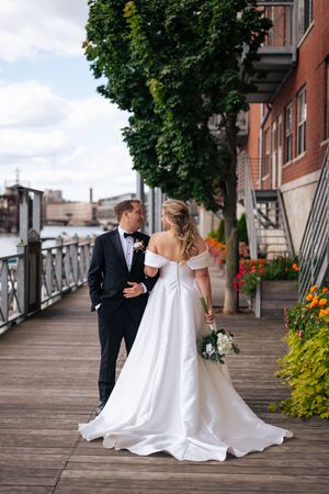 A couple in wedding attire walking along a waterfront boardwalk with trees and flowers in the background.