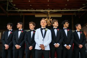 Groom in white tuxedo jacket stands with six groomsmen in black tuxedos and bow ties with red boutonnieres.