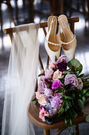 Badgley Mischka bridal heels with pearl straps rest on a wooden chair beside a lush purple and pink wedding bouquet.