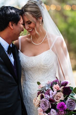 Bride in beaded gown and veil holding purple bouquet smiles with groom in tuxedo at outdoor wedding.