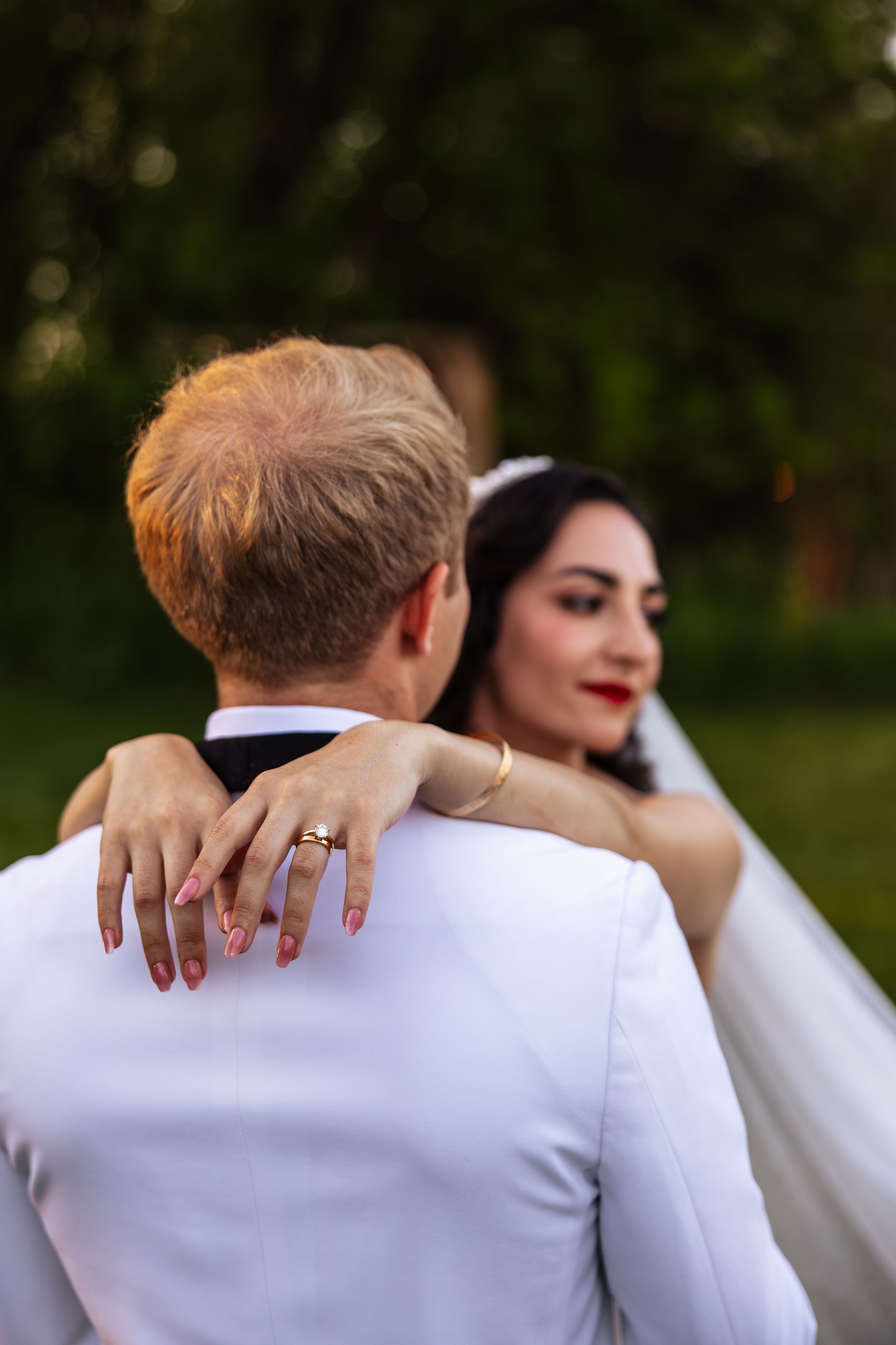 Bride with red nails and gold ring embraces groom in white suit outdoors at wedding ceremony.
