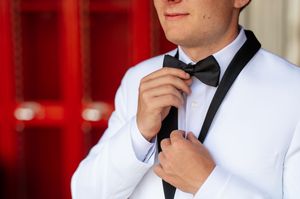 Man in white tuxedo jacket adjusting black bow tie, formal wedding attire against red background.
