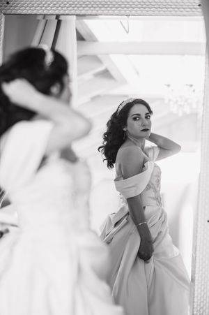 Black and white photo of a bride in an off-shoulder wedding gown adjusting her hair while gazing into a mirror.