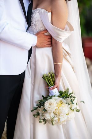 Bride in off-shoulder white gown holds bouquet of white roses and greenery while groom embraces her waist.
