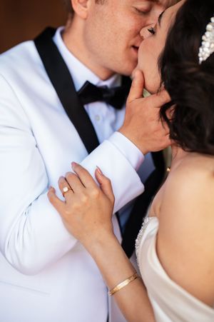 A bride and groom share a romantic kiss, with the bride wearing a gold bracelet and engagement ring on her hand.