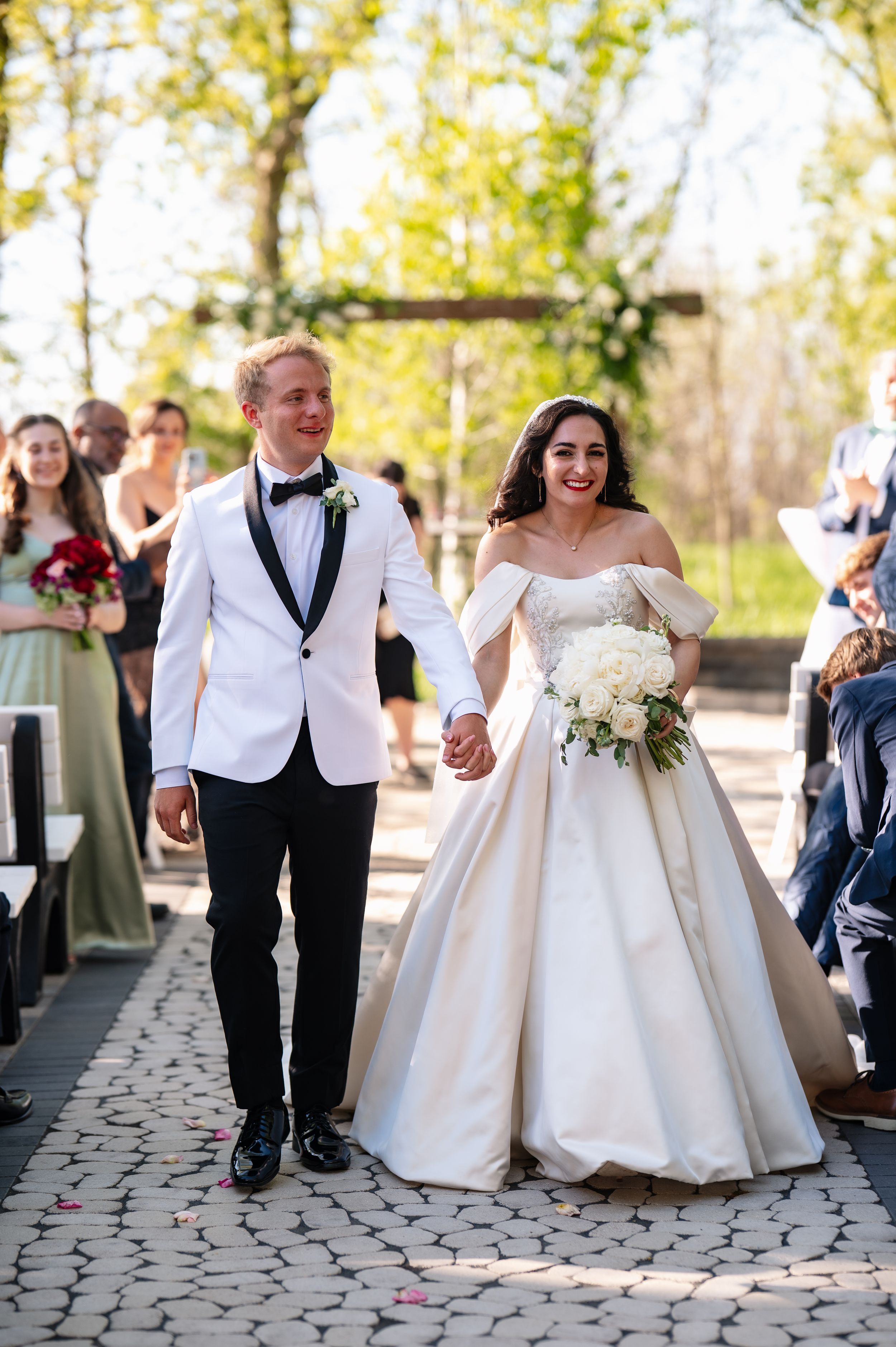 Smiling newlyweds walk down aisle, groom in white tuxedo, bride in off-shoulder ball gown holding white bouquet.