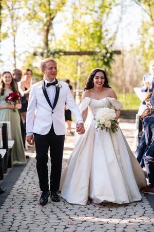 Smiling newlyweds walk down aisle, groom in white tuxedo, bride in off-shoulder ball gown holding white bouquet.