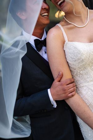 Bride and groom laughing together, she wears a beaded dress and pearl necklace, he wears a black tuxedo with bow tie.