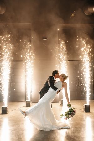 Bride and groom share a romantic dip kiss surrounded by sparkling indoor fireworks fountains during their wedding reception.