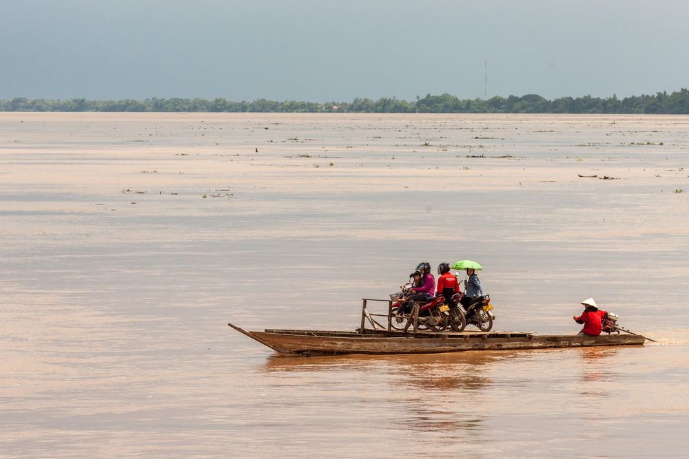 Small wooden boats navigate across a wide expanse of muddy river water during low tide conditions.