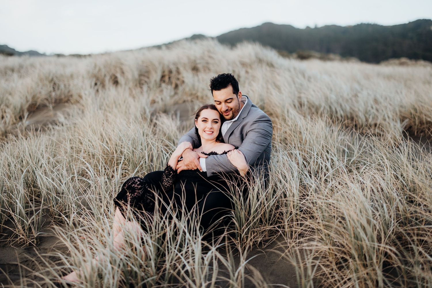 A couple embraces tenderly in a field of tall dry grass as the sun sets behind mountain peaks.