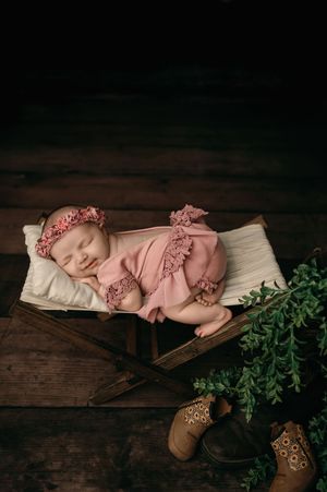 A newborn in pink ruffled outfit sleeps peacefully on white cushion in vintage wooden box with greenery and boots nearby.
