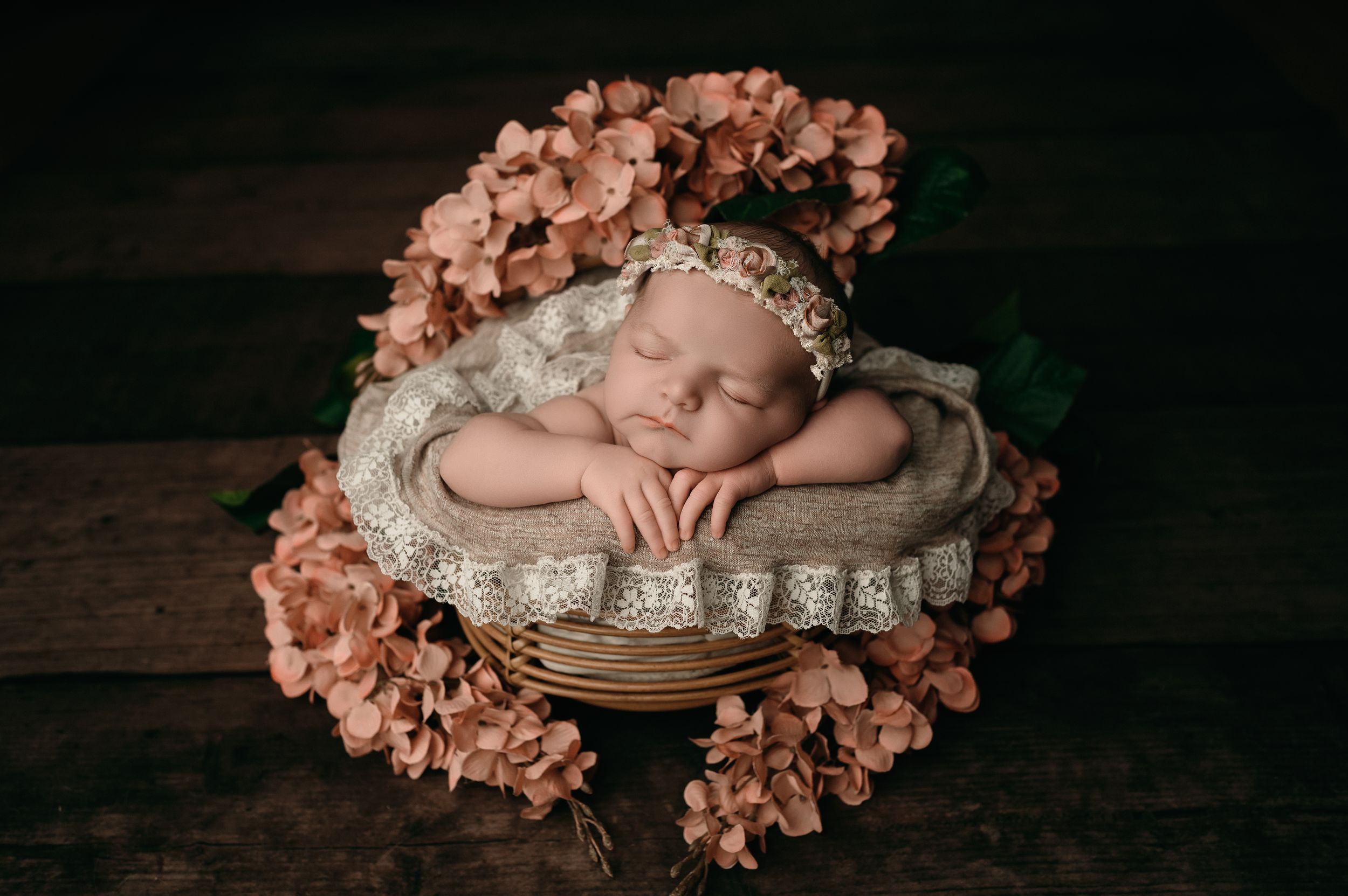 A sleeping newborn in a woven basket surrounded by soft peach-colored floral arrangements on a rustic wooden surface.