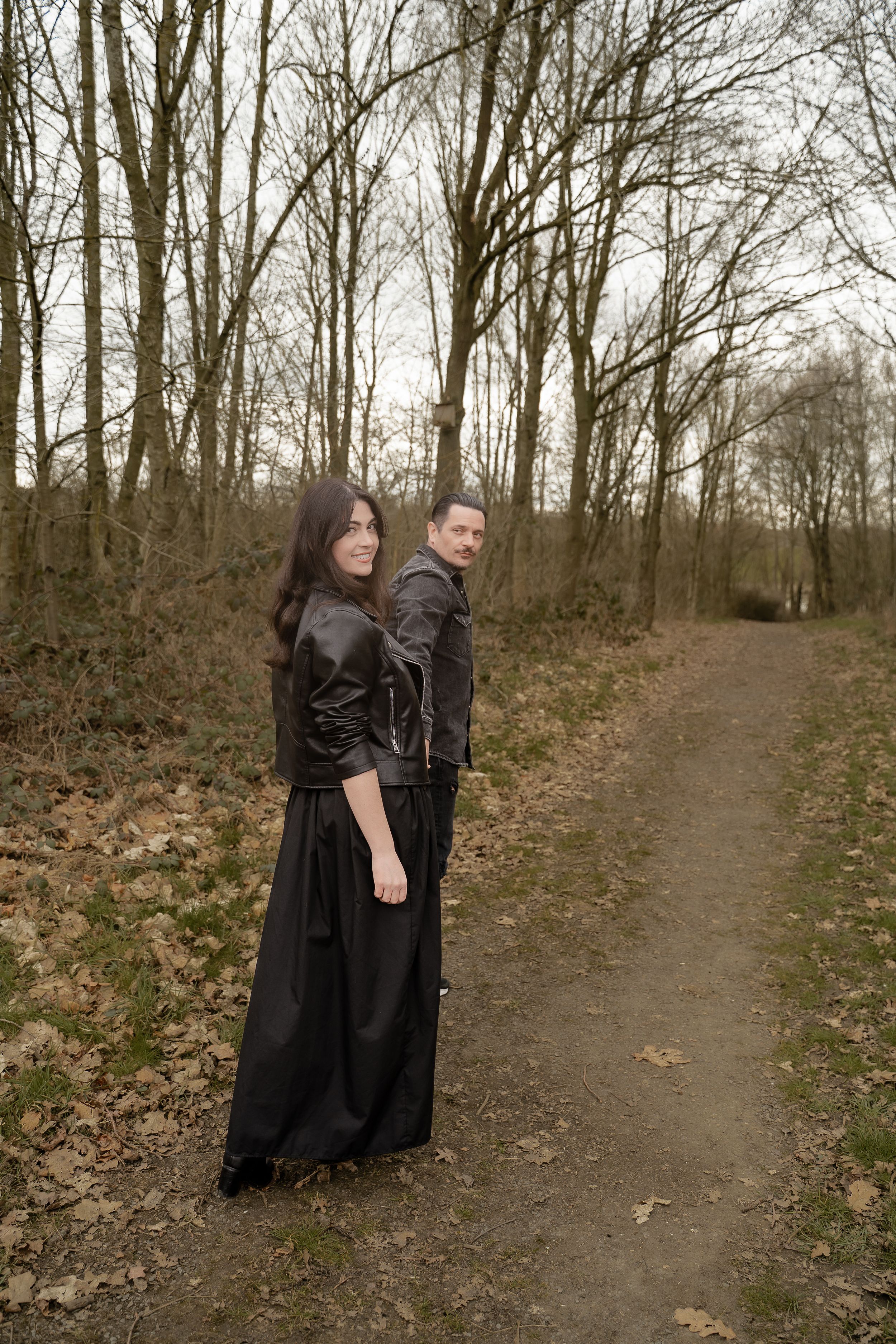 Two people in dark clothing stand together on a forest path during autumn with bare trees in the background.
