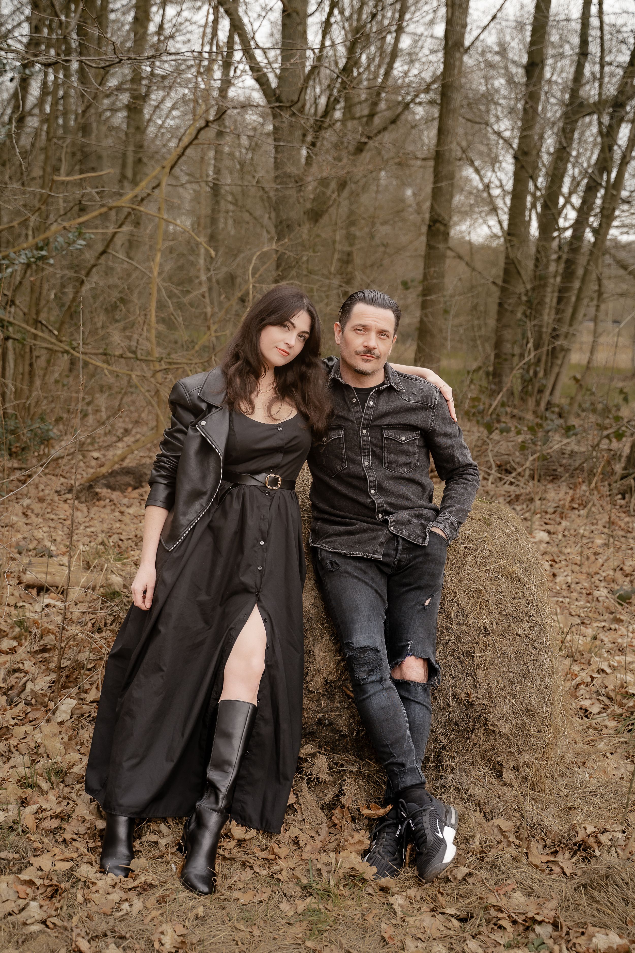 A couple in dark clothing sits casually together on a wooded hillside during a photo session.