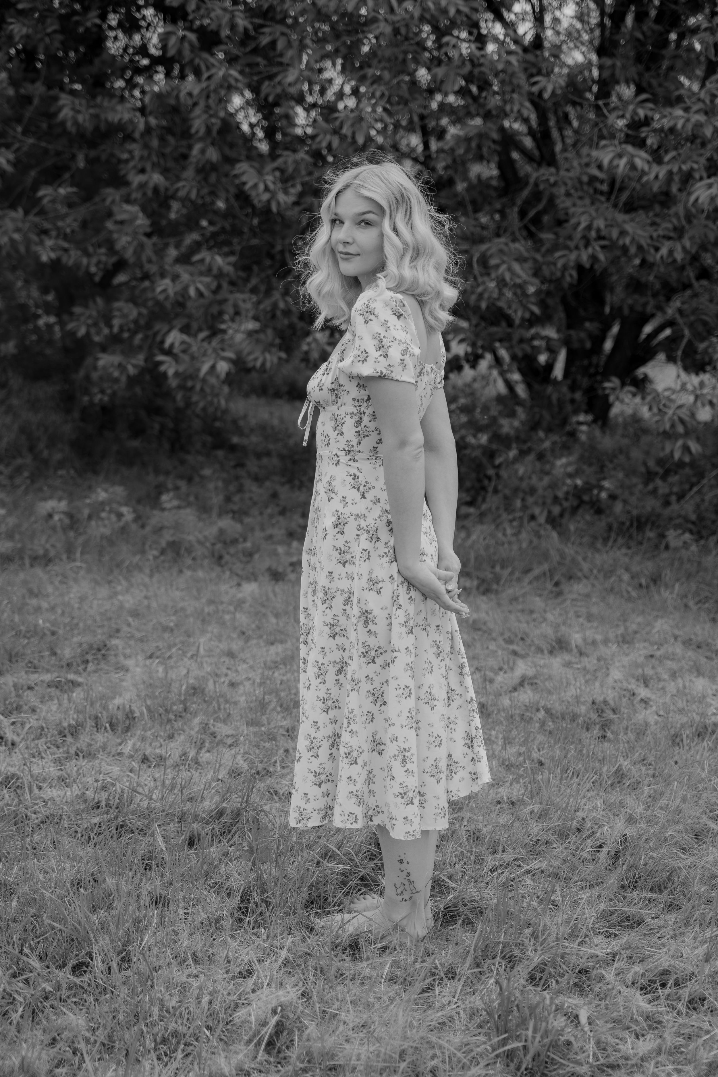 Black and white photograph of a person in a floral dress standing in a grassy outdoor setting.