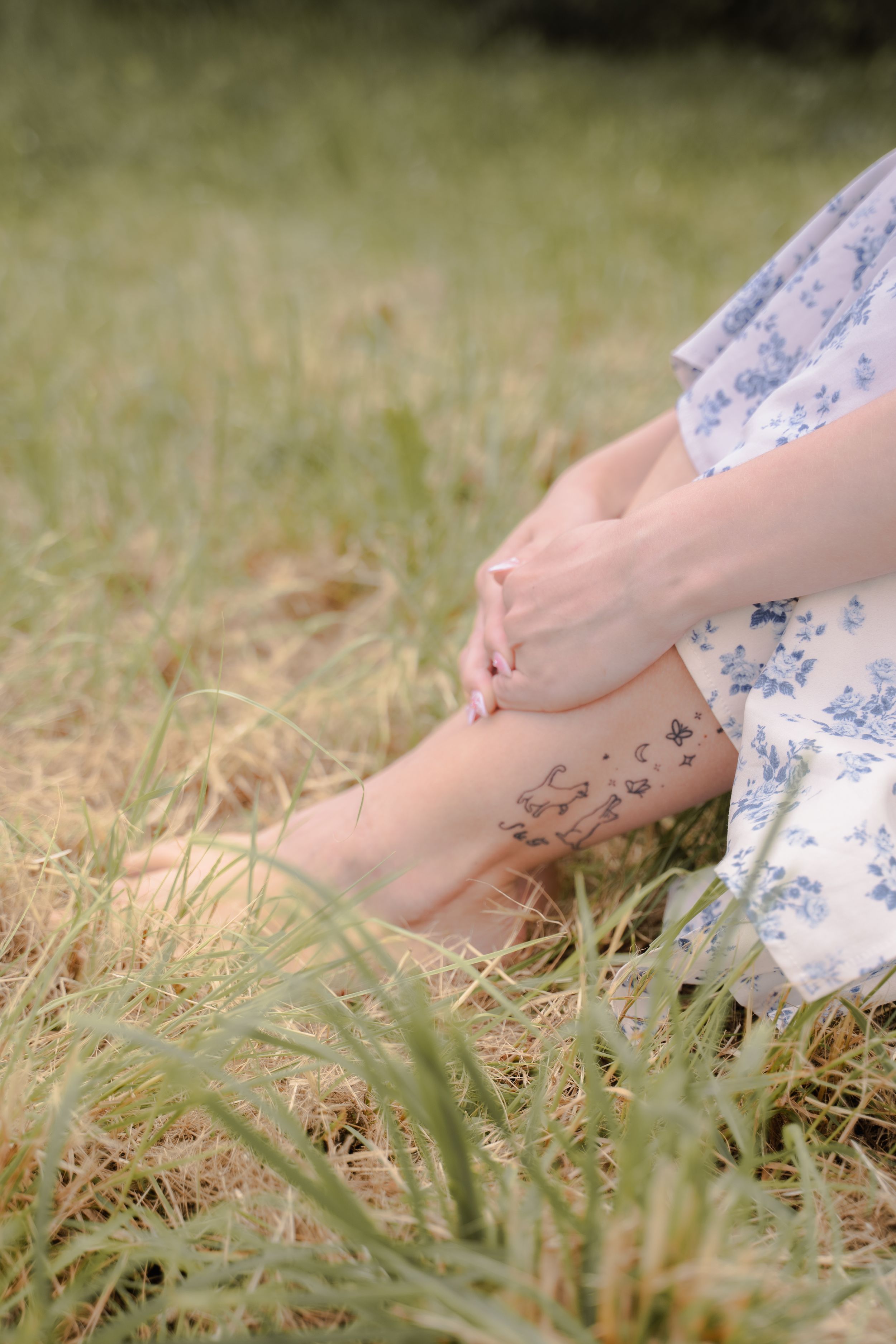 A hand in a white floral dress gently touches tall grass in a meadow during golden hour.