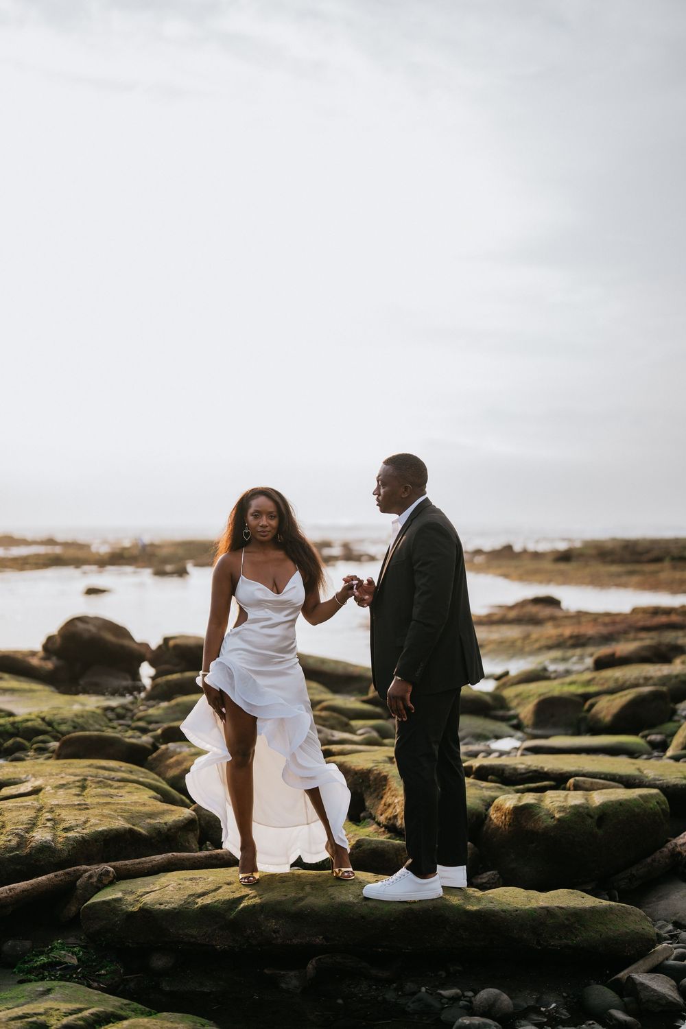 Wedding couple in formal attire stands on mossy coastal rocks at sunset.