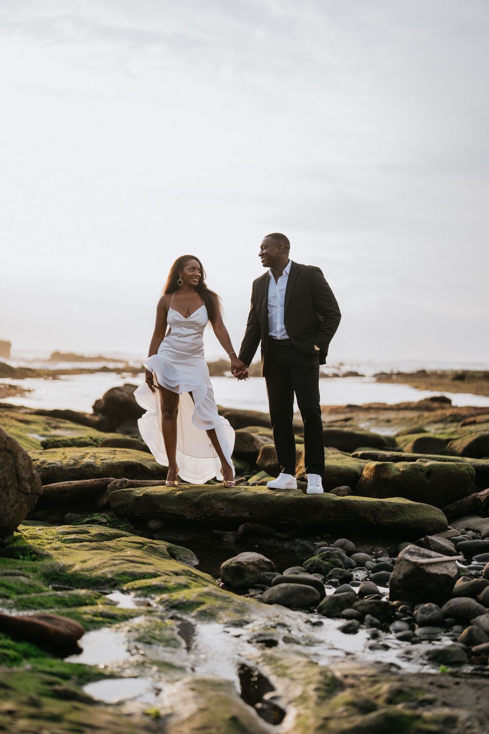 A couple in formal attire poses intimately on mossy coastal rocks during a seaside photoshoot.