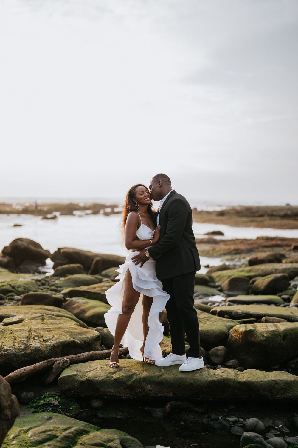 A tender moment captured between a couple sitting on moss-covered rocks by the ocean.