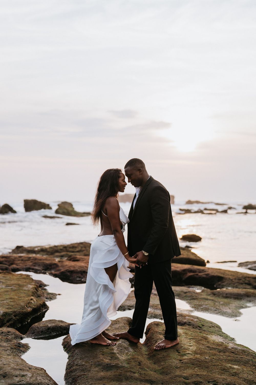 A couple in formal wear shares a romantic moment on coastal rocks at dusk.