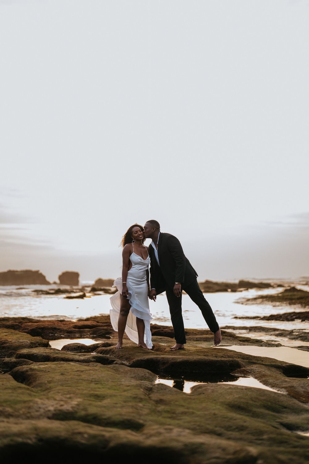 A couple in formal attire poses dramatically on rocky coastal terrain.
