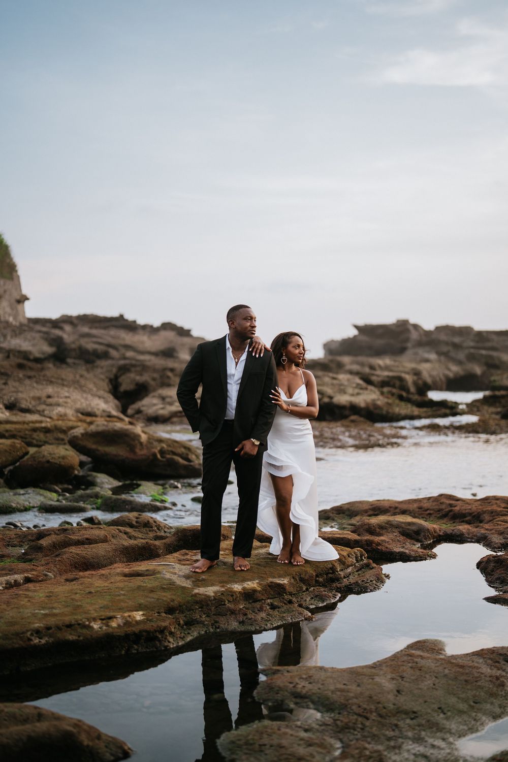 A couple in wedding attire explores tidal pools among coastal rocks.