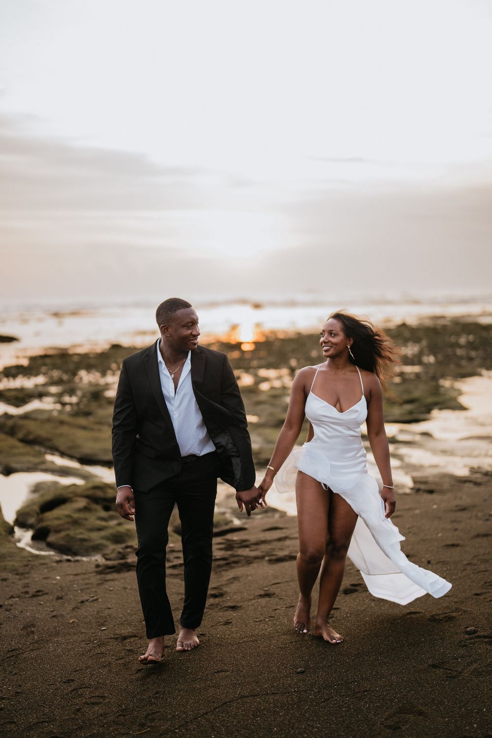 A couple playfully walking along a dark sandy beach at sunset.