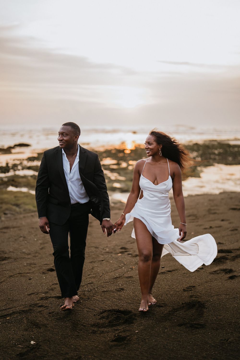 A romantic couple strolling on a beach with flowing white dress and black suit.