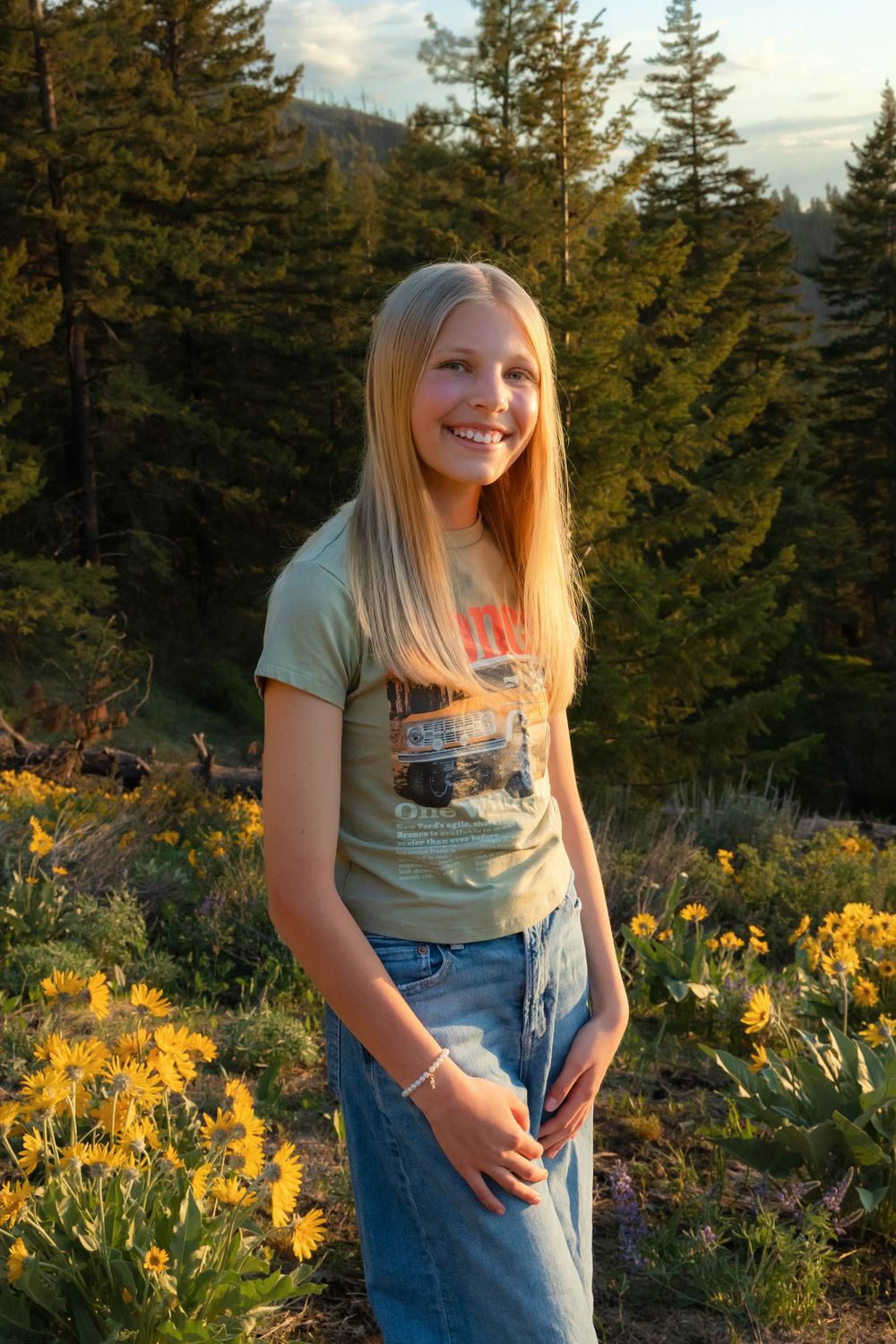 Someone in casual denim and t-shirt poses among blooming yellow flowers with evergreen trees behind.