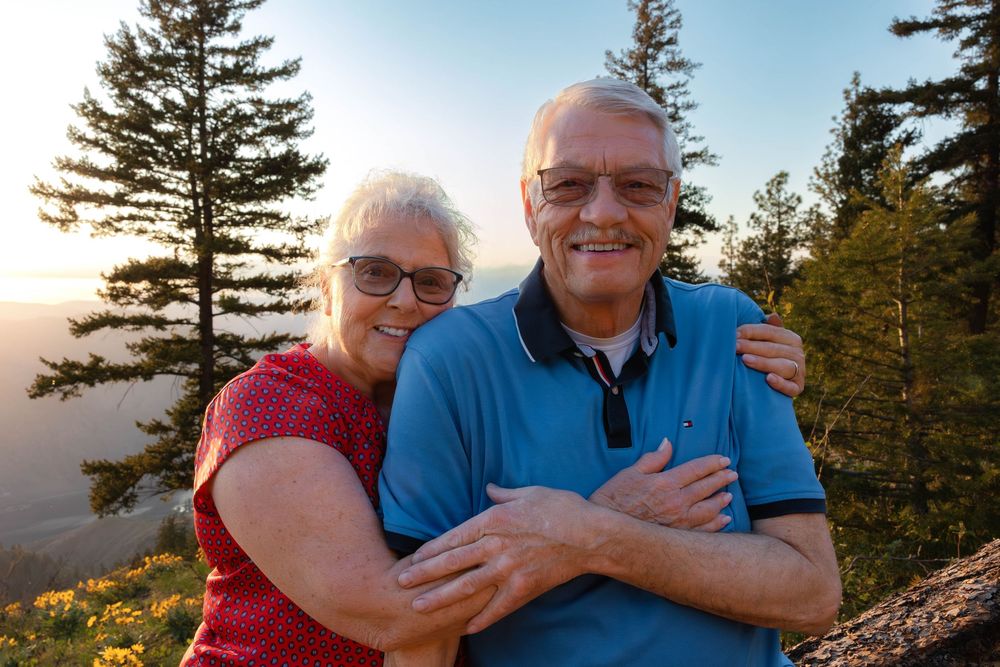Two people embrace while enjoying a sunset among evergreen trees.