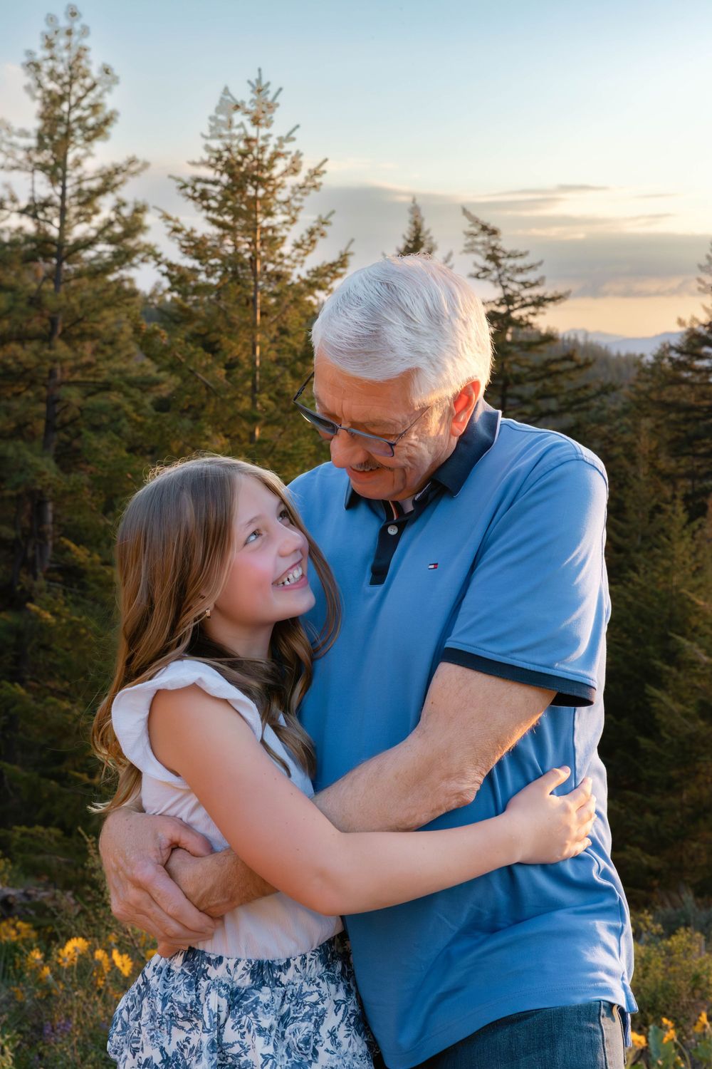 Tender family moments captured during sunset with evergreen trees and yellow flowers in background.