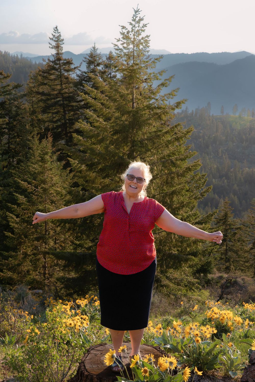 Person in red shirt stands with arms outstretched on mountain overlook surrounded by evergreens and wildflowers.
