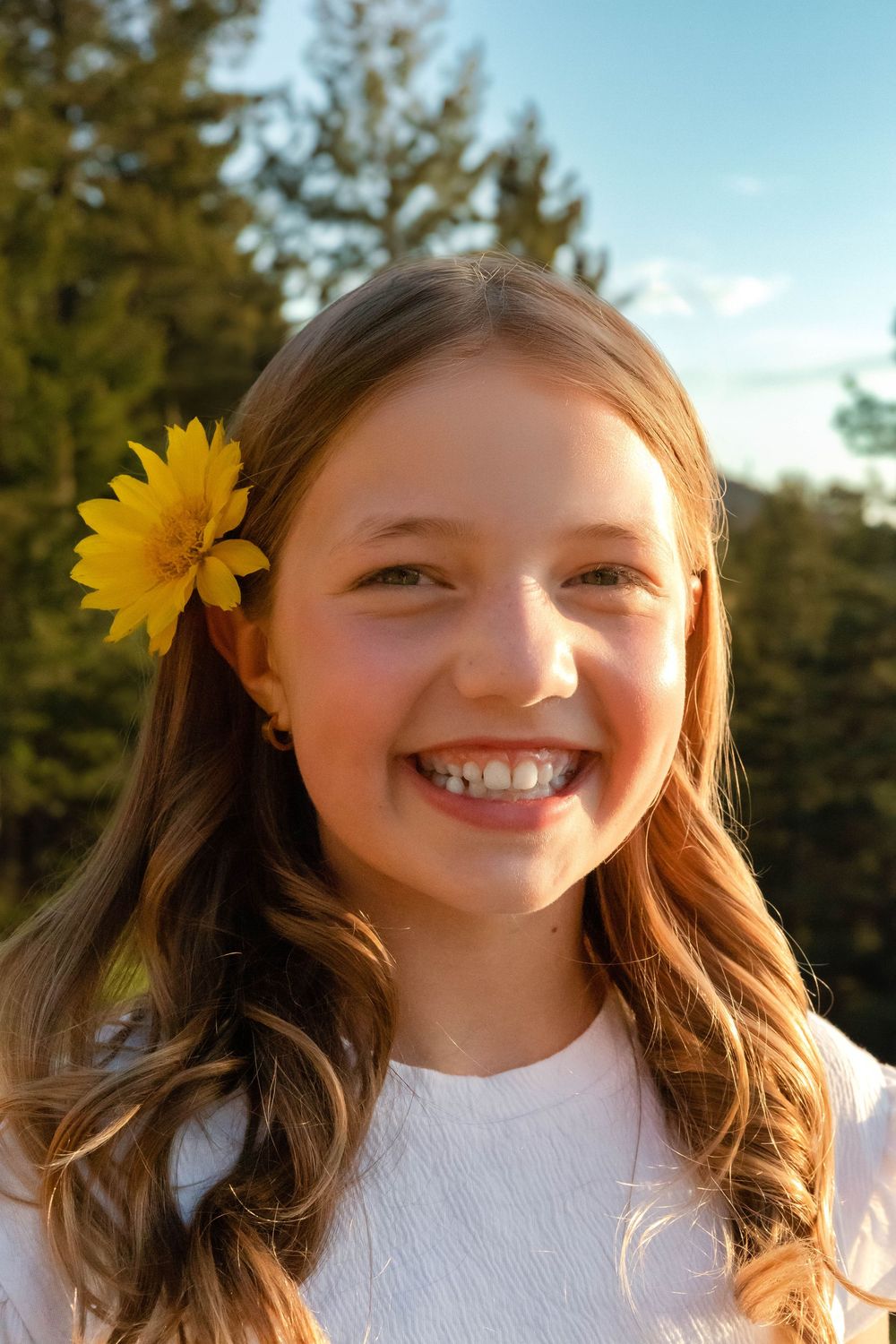 Young person wearing white top with yellow flower in hair smiles at camera against natural outdoor setting.