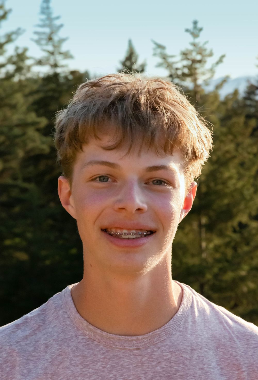 Portrait of a young person smiling outdoors with evergreen trees in the background.