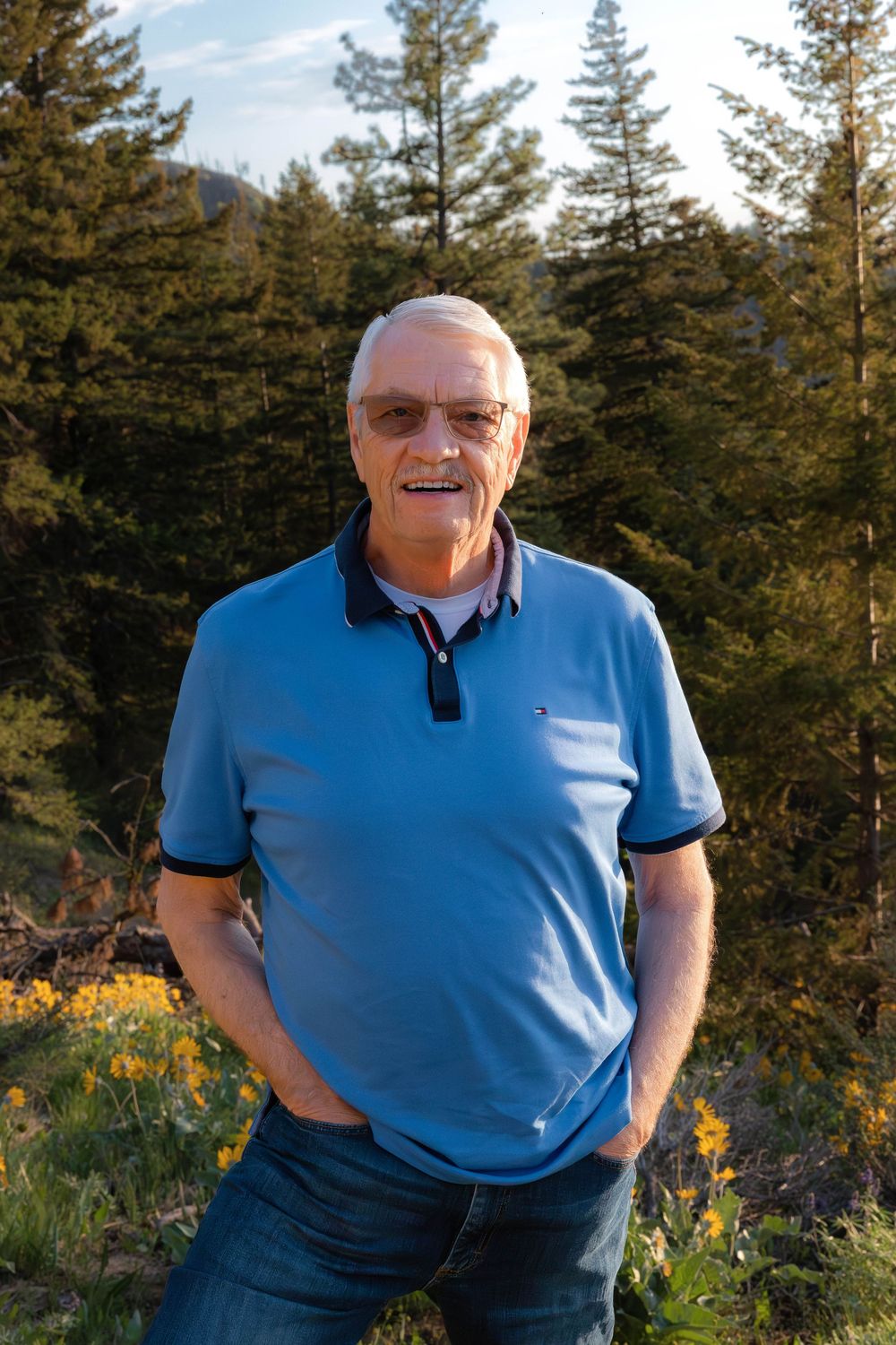 Person in blue polo shirt smiling outdoors with evergreen forest backdrop.