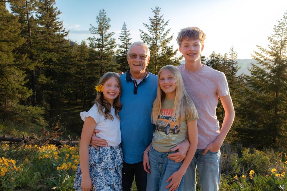 Family group portrait outdoors with evergreen trees and mountain scenery in background.