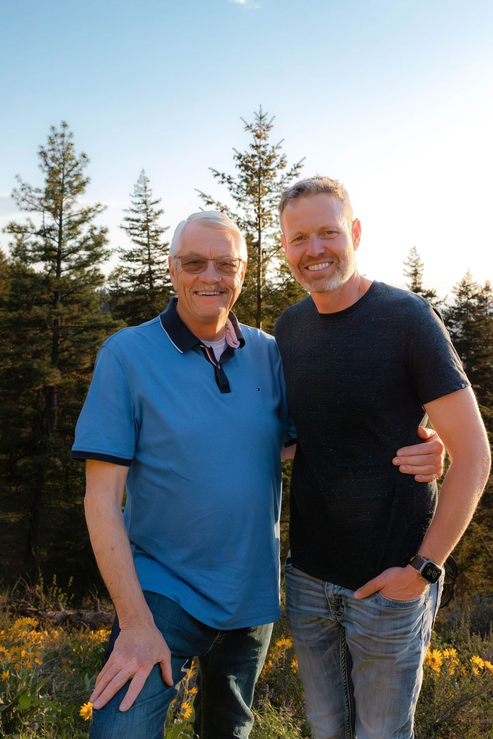 Two people in casual attire share laughs while posing against mountain landscape with pine trees.