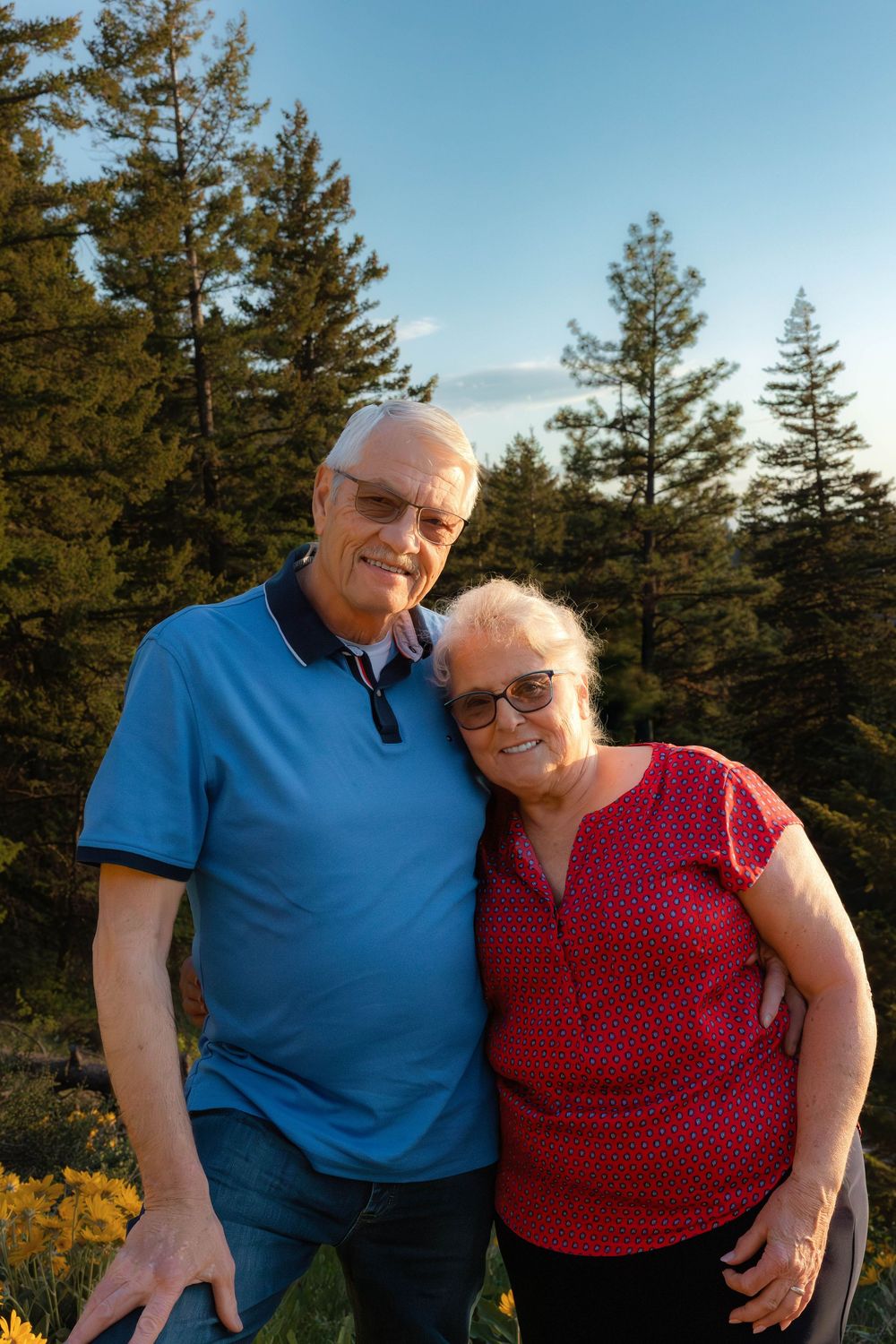 Couple in blue and red shirts embrace while posing outdoors against evergreen forest backdrop.