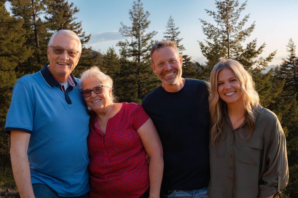 A group of four people smiling together outdoors with evergreen trees in the background during sunset.