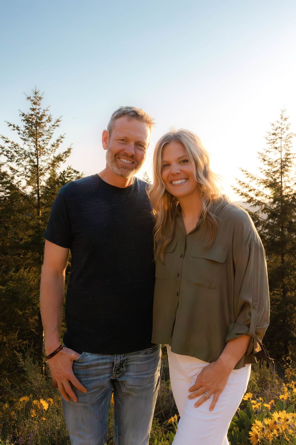 Couple in casual wear shares intimate moment while posing in natural setting with sunset lighting.