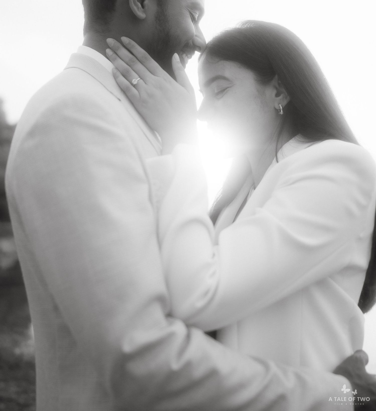 A tender black and white portrait of a couple in white clothing embracing in sunlit setting.