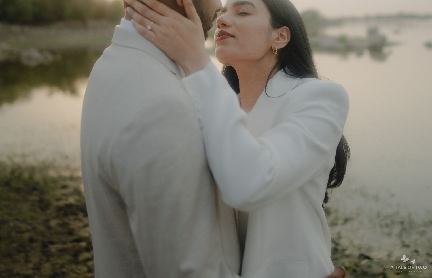 A romantic couple in white outfits embraces intimately during an outdoor photoshoot at golden hour.