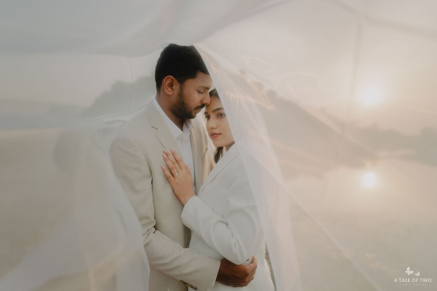 A romantic couple in cream-colored outfits poses together against a dreamy sunlit background.