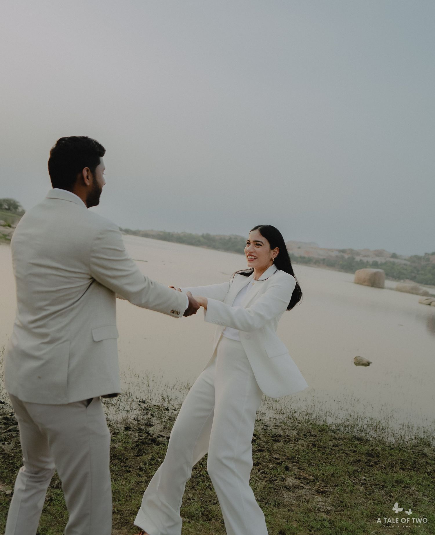 A couple in white and beige outfits playfully dance together on a grassy hilltop during golden hour.