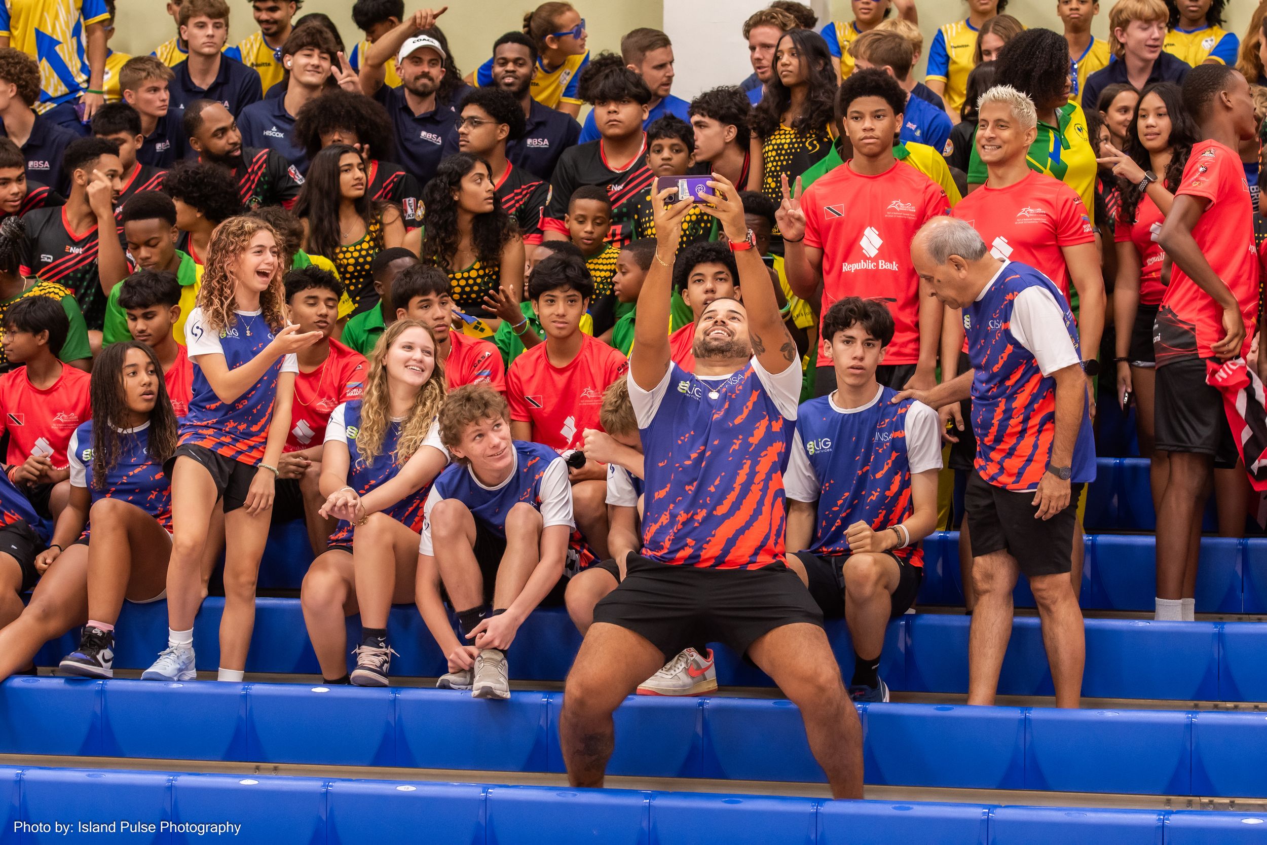 Diverse group of young athletes sitting on bleachers at an indoor sports event, wearing colorful team uniforms.