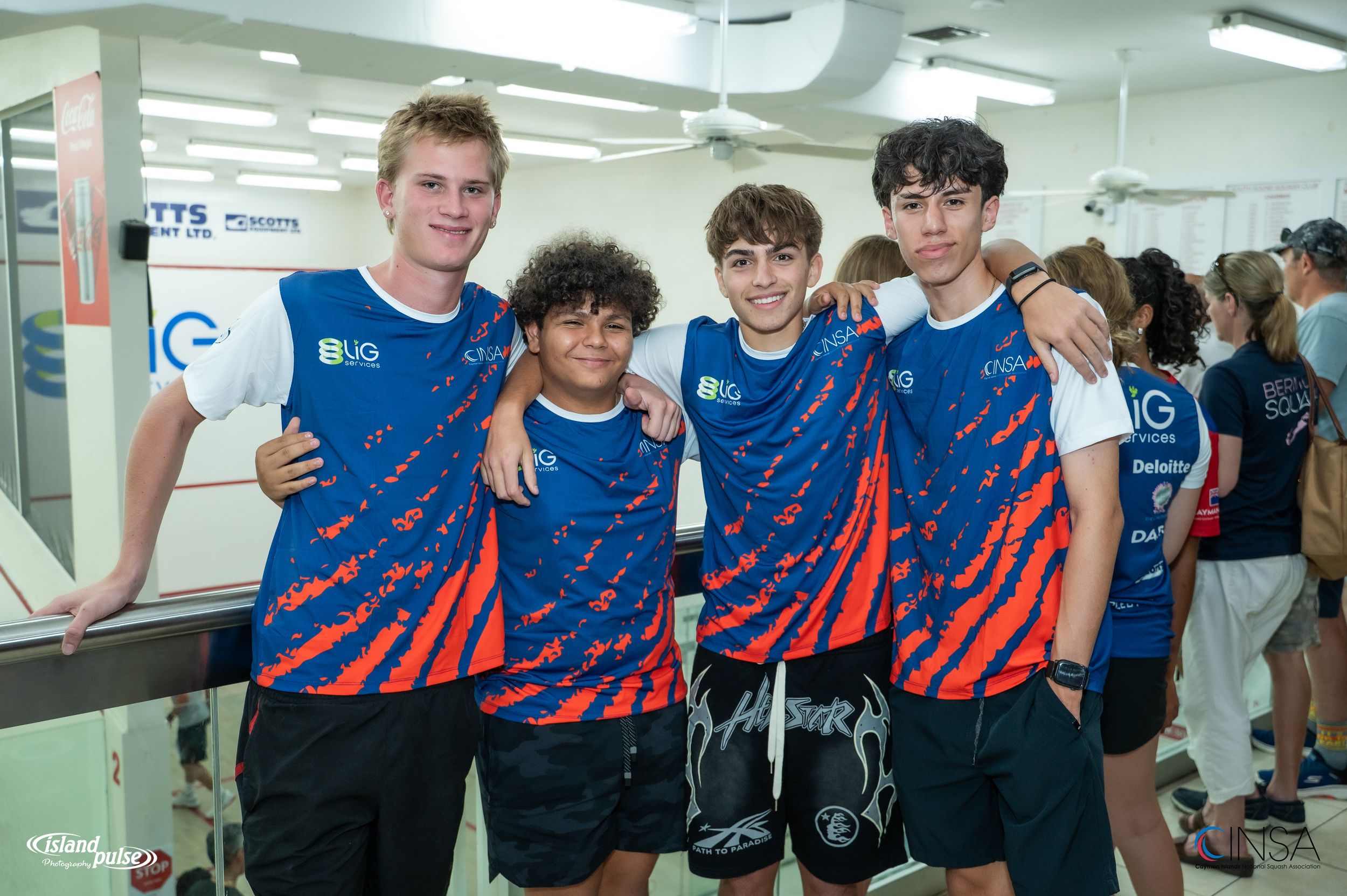 Four teenage boys in matching blue and red jerseys pose together smiling at an indoor sports facility.